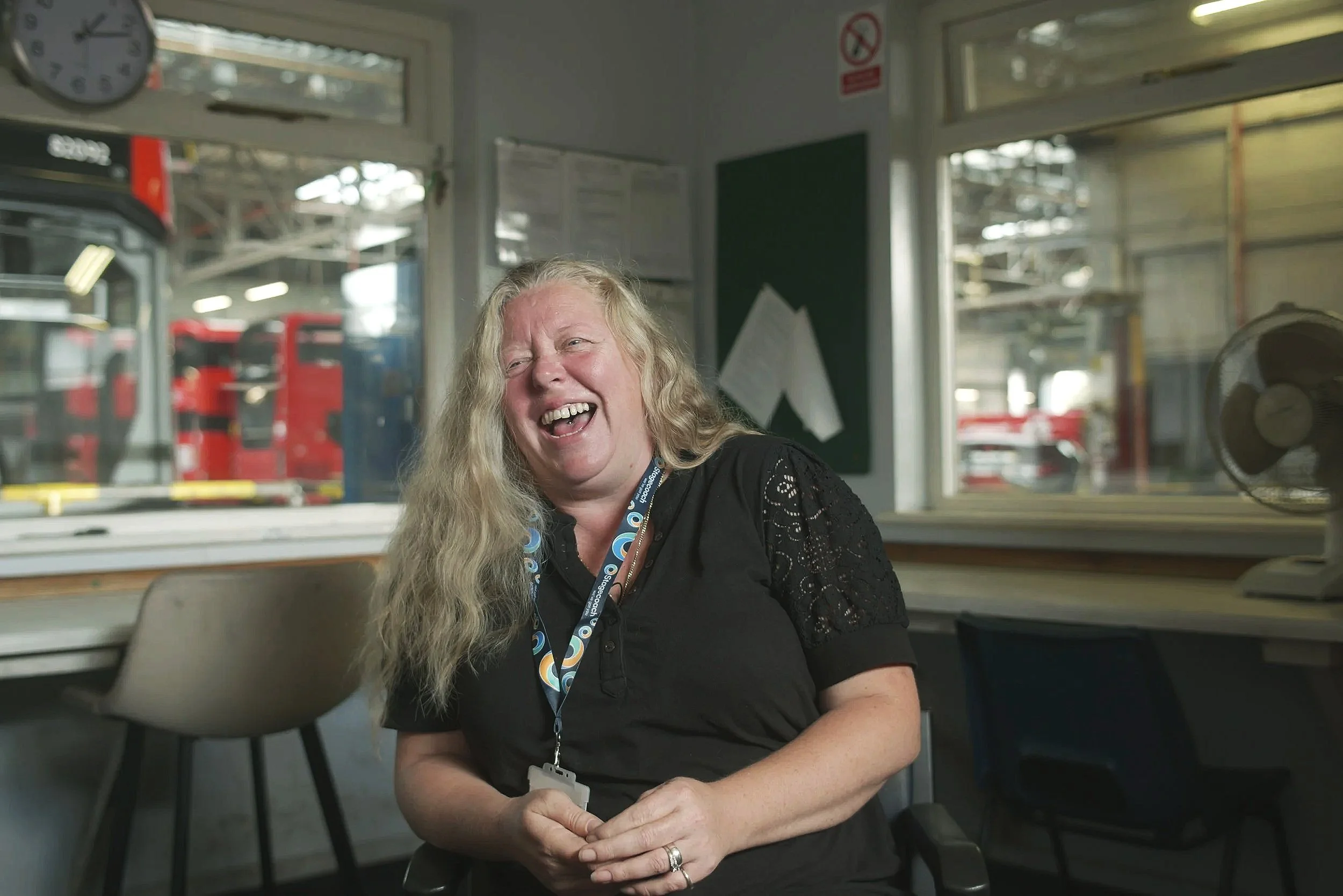 A woman with long blonde hair, wearing a black shirt with lace sleeves, is sitting in an office or control room, laughing with her eyes closed. There are windows behind her showing a garage or station with red vehicles.