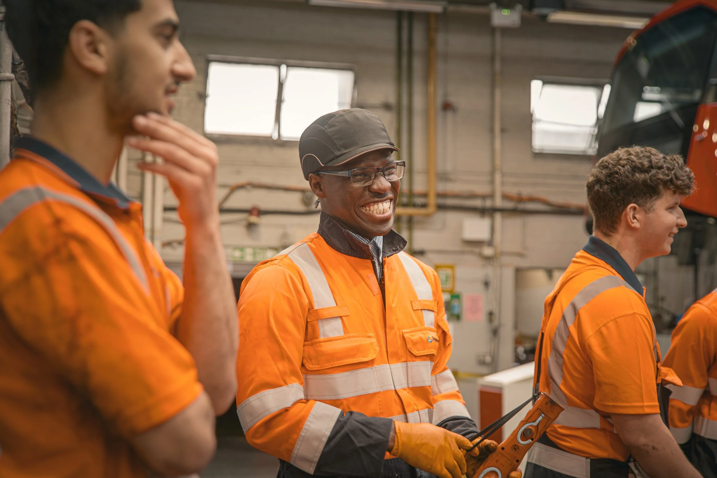 Group of workers in orange safety uniforms, smiling and talking inside a warehouse or industrial facility.