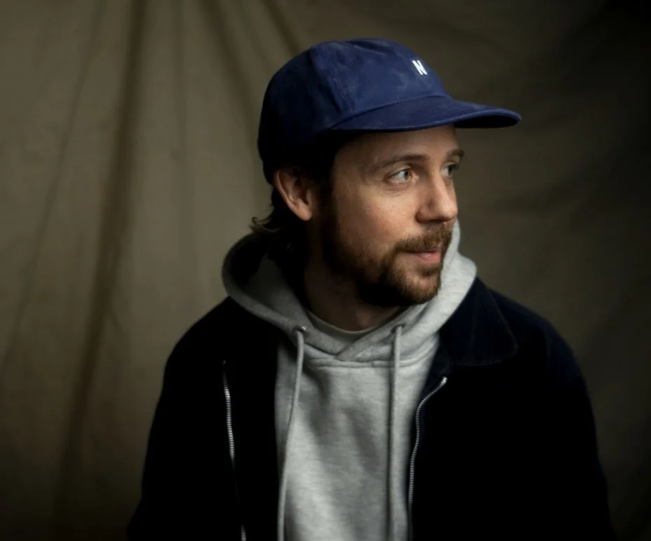 A man with a beard and mustache wearing a navy baseball cap with 'N' on it, a gray hoodie, and a dark jacket, looking to the side against a plain beige background.