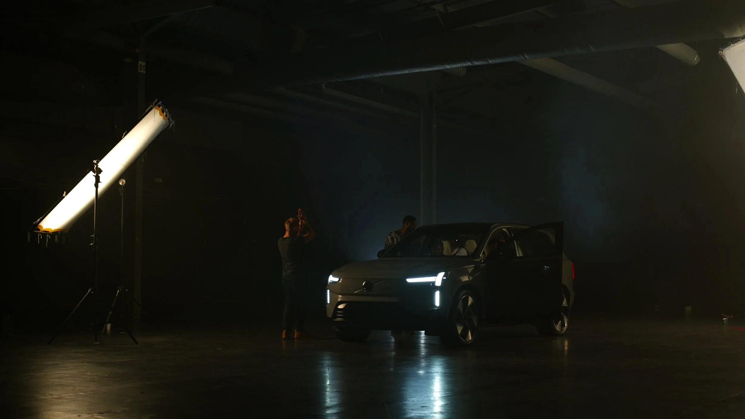 Behind the scenes of a car photo shoot in a dark studio with professional lighting equipment, including a large softbox. A black vehicle is positioned in the center, with people preparing and filming around it.