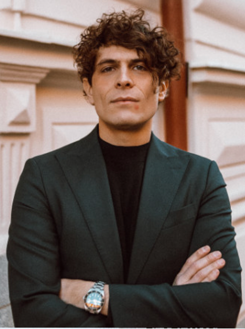 A young man with curly hair, wearing a black turtleneck and green blazer, standing outdoors with arms crossed in front of beige siding.