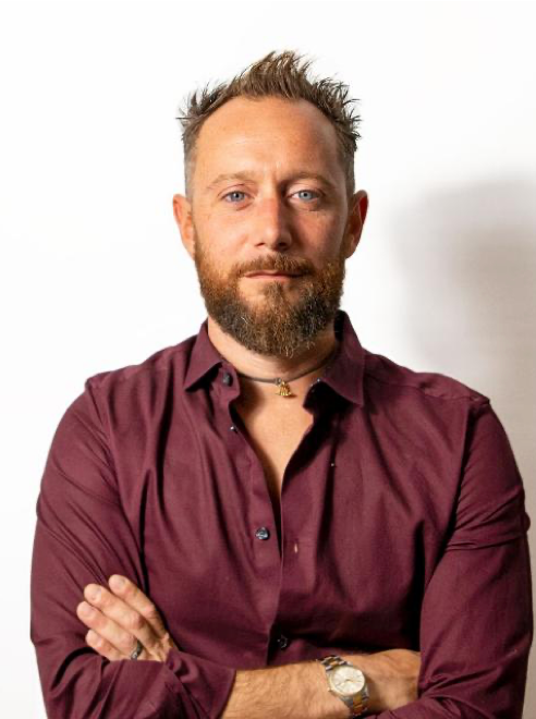 A man with a beard and short, spiked hair wearing a maroon button-up shirt with sleeves rolled up, standing with arms crossed against a plain white background.