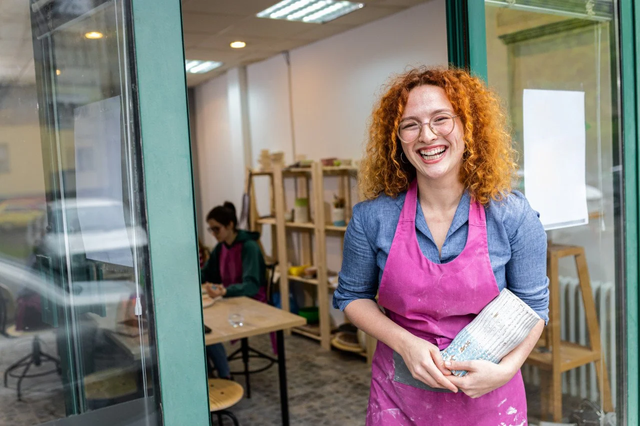 A woman with curly red hair, glasses, and wearing a pink apron over a blue shirt is smiling and holding a ceramic object inside a shop or studio, with another person working at a table in the background.