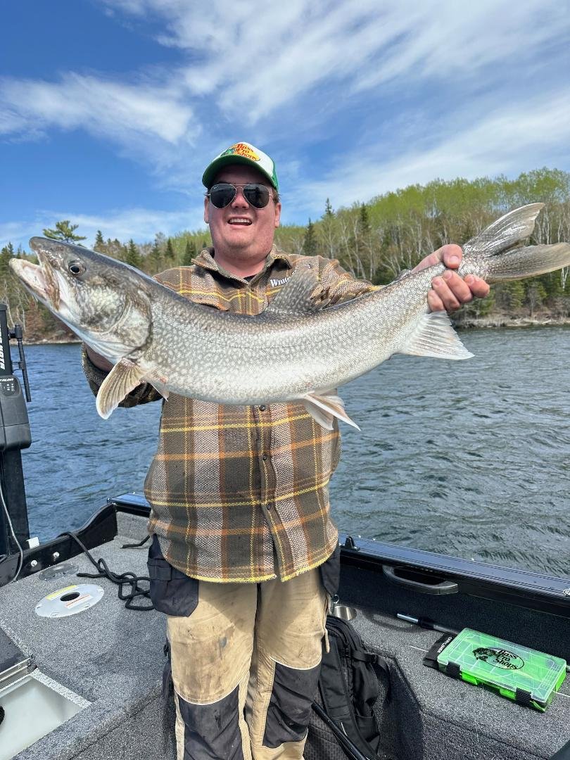 Person holding a large fish on a boat with trees and water in the background.