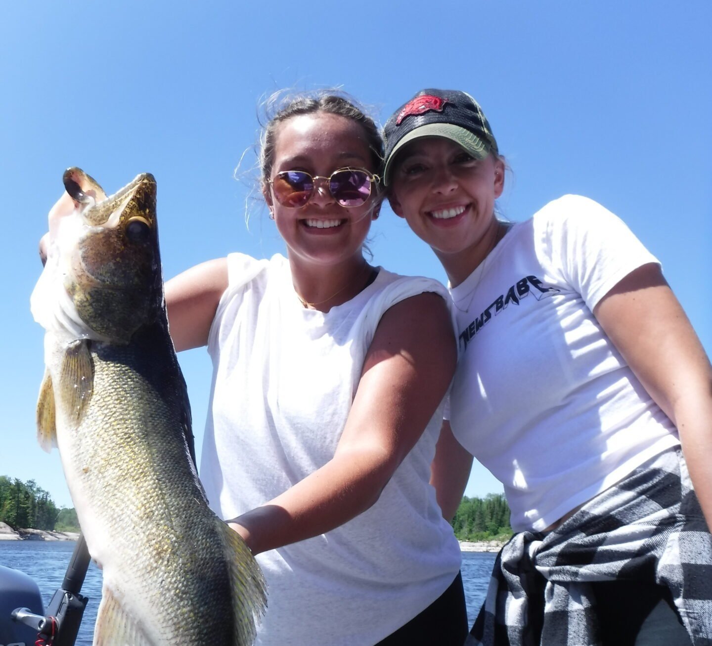 Two women smiling, holding a large fish, outdoors with a clear blue sky.