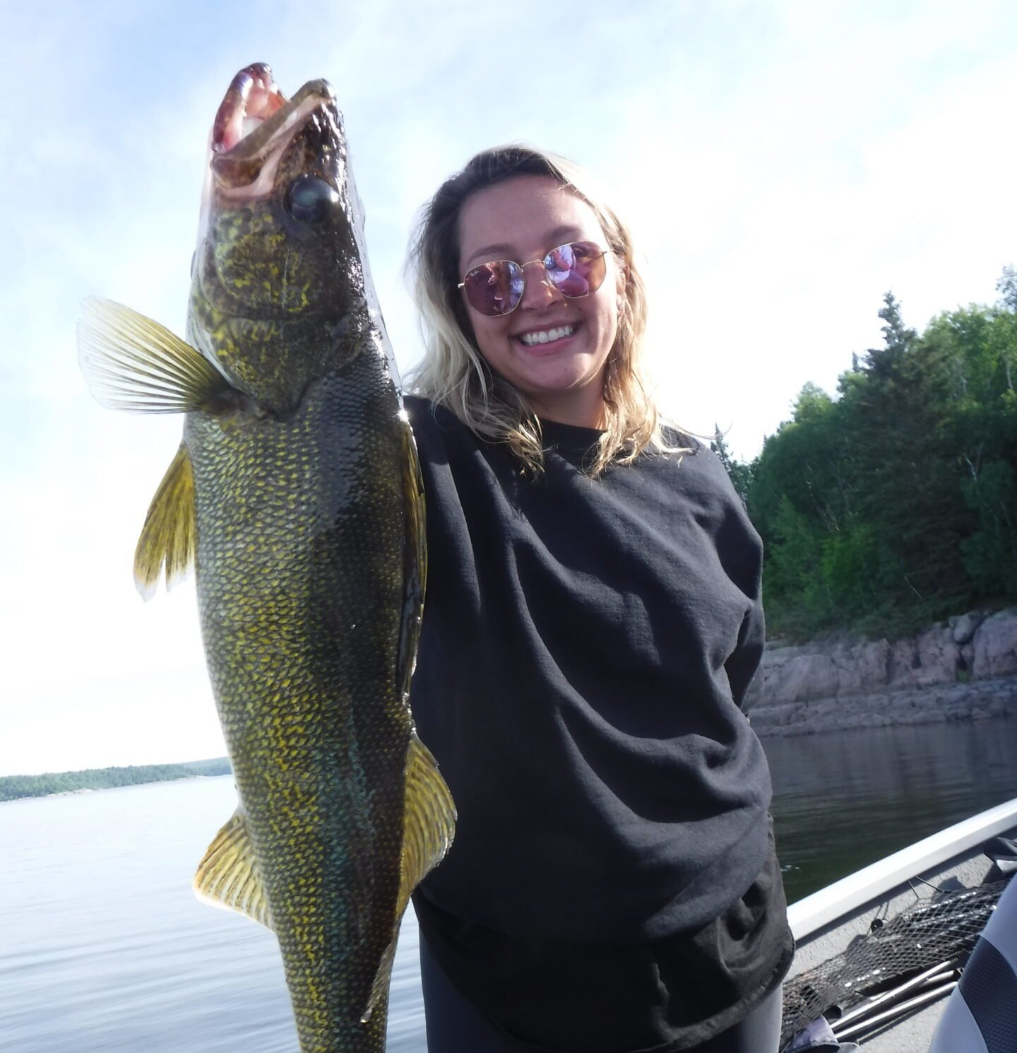Person holding a large fish on a boat with water and trees in the background.