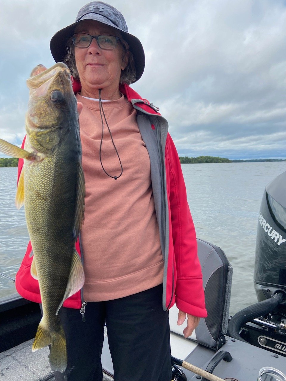 Person holding a fish on a boat, wearing a red jacket and hat, with water and cloudy sky in the background.