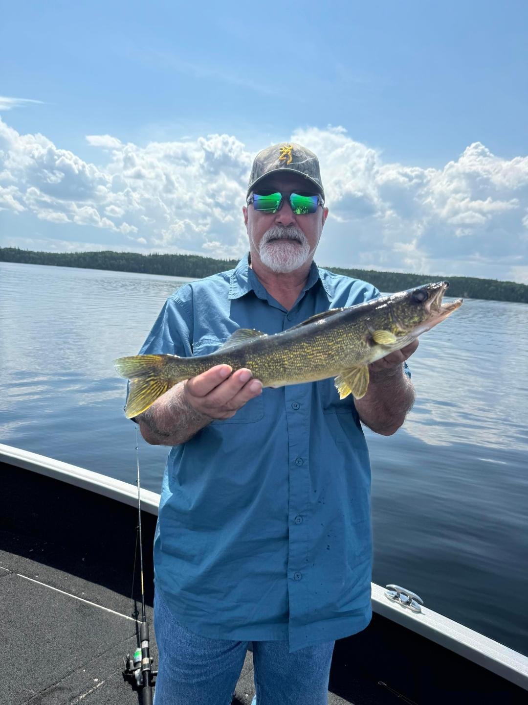 Man holding a fish on a boat, wearing sunglasses and a cap, with blue sky and water in the background.