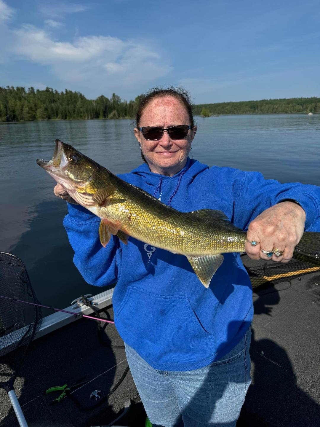 Person holding a large fish on a boat with a lake and forest in the background.