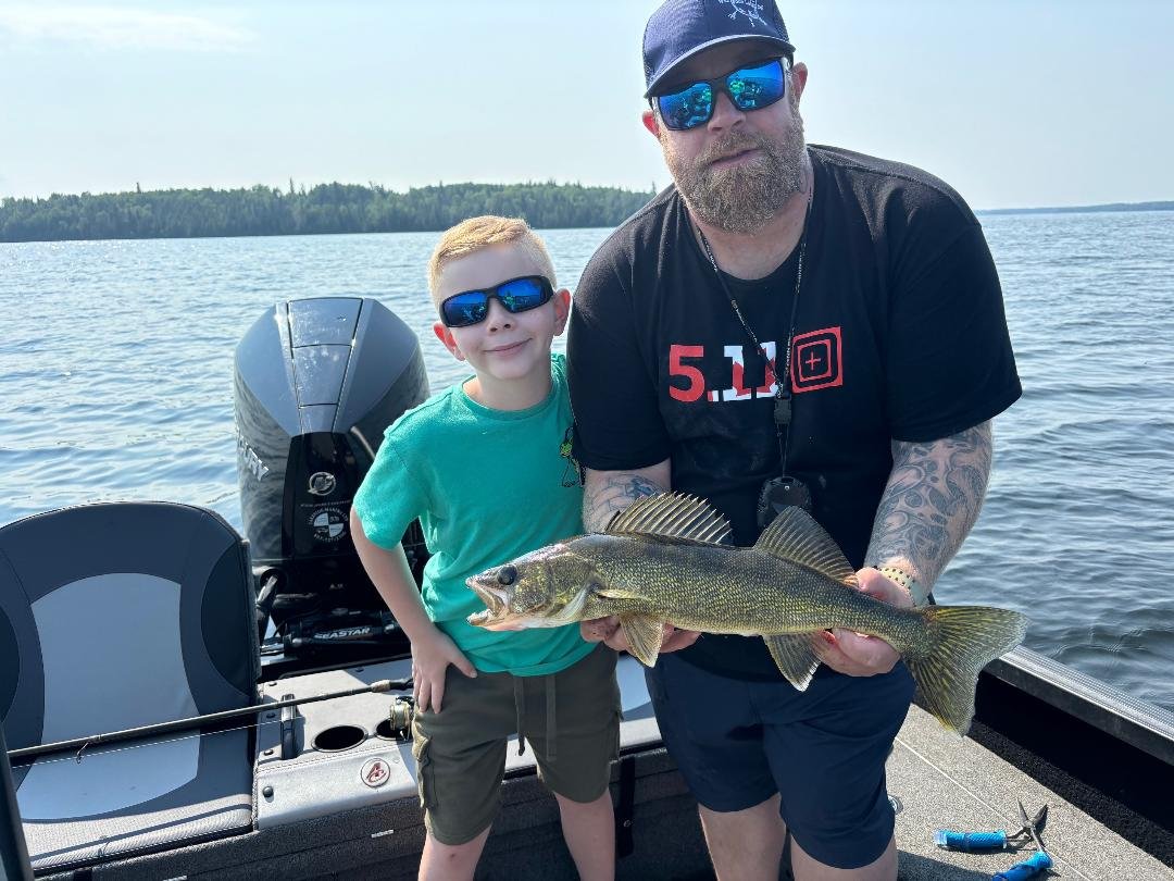 Father and son fishing on a boat, holding a fish, with a lake and trees in the background.