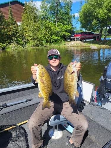 Person sitting in a boat holding two large fish on a river with trees in the background.
