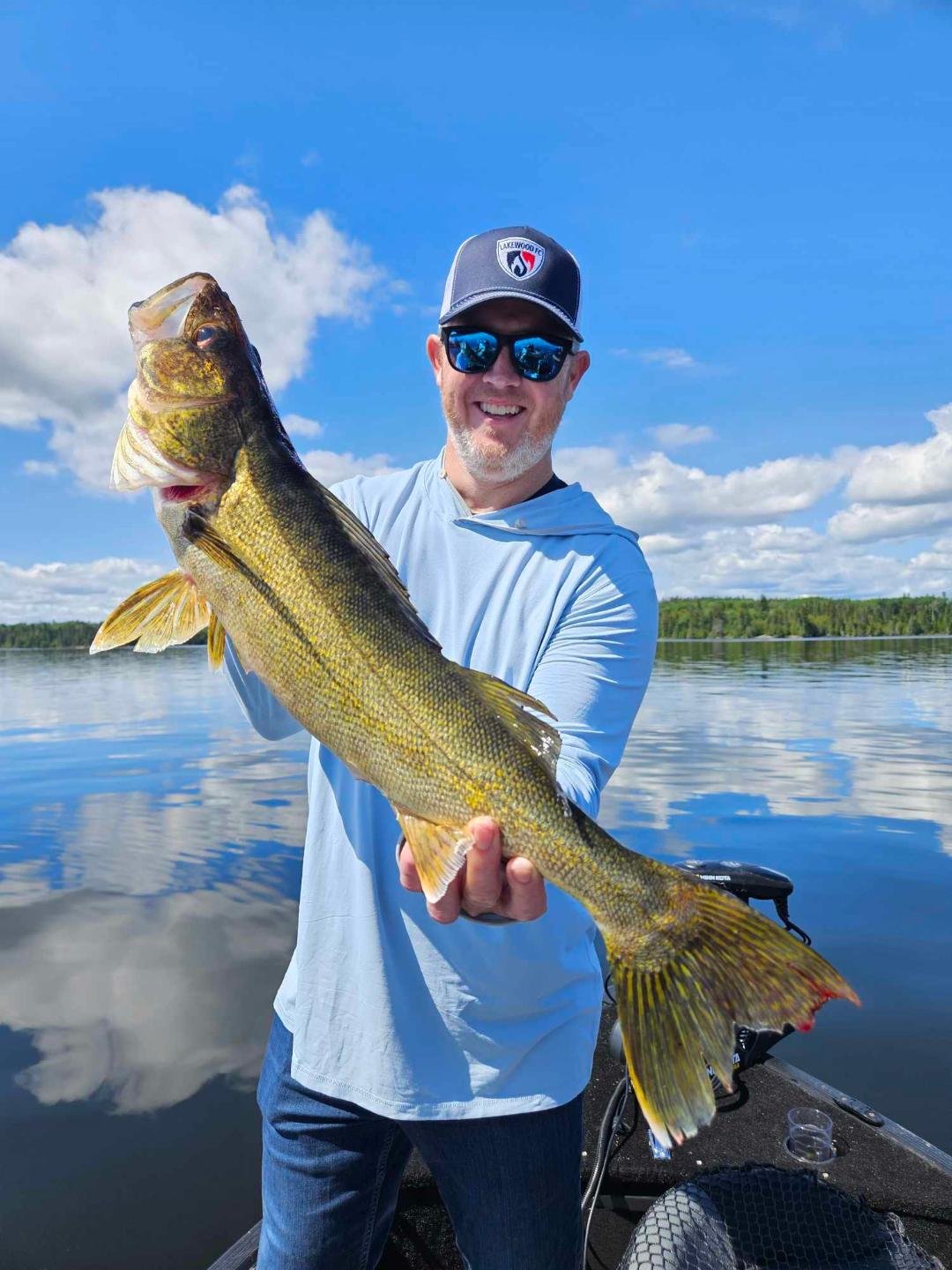 Person holding a large fish on a lake, wearing a baseball cap and sunglasses, with a blue sky and clouds in the background.