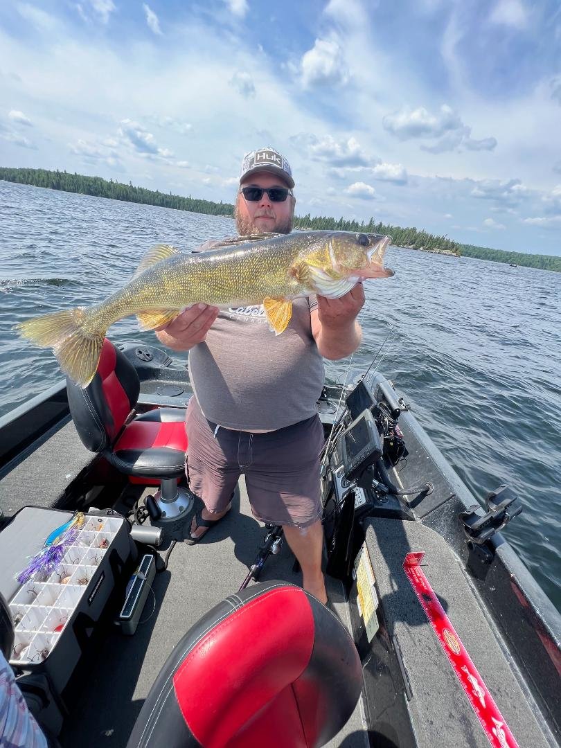 Man holding a large fish on a boat in a lake with fishing gear visible.
