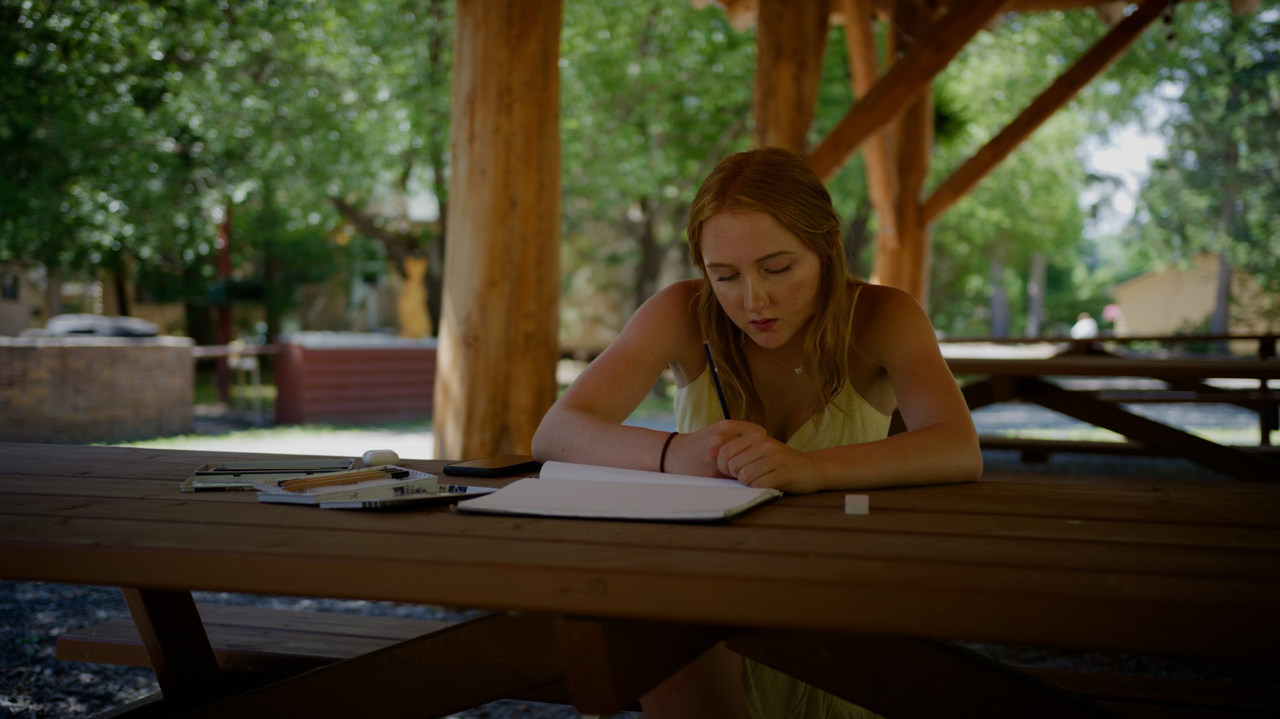 Person writing in a notebook at a wooden picnic table in an outdoor setting.