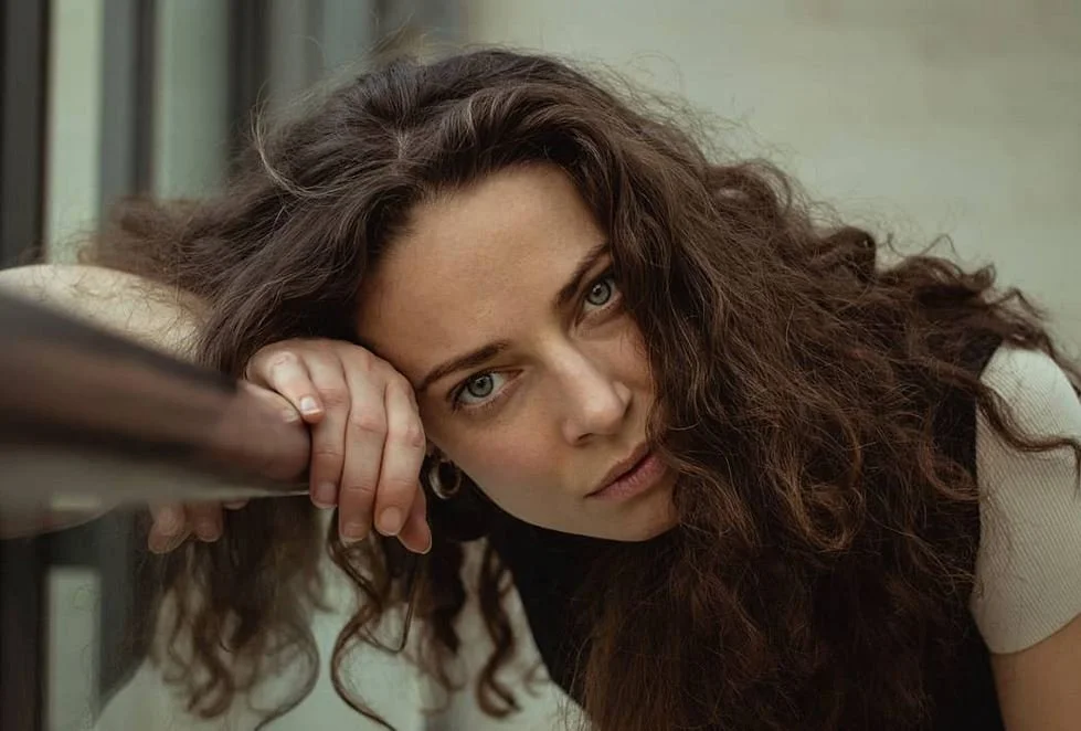 Kvindelig person med langt, krøllet brunt hår, hvide øjne, hvide ører og let bleg hud, hviler sit hoved på en hånd mod en væg i en indendørs indstilling.