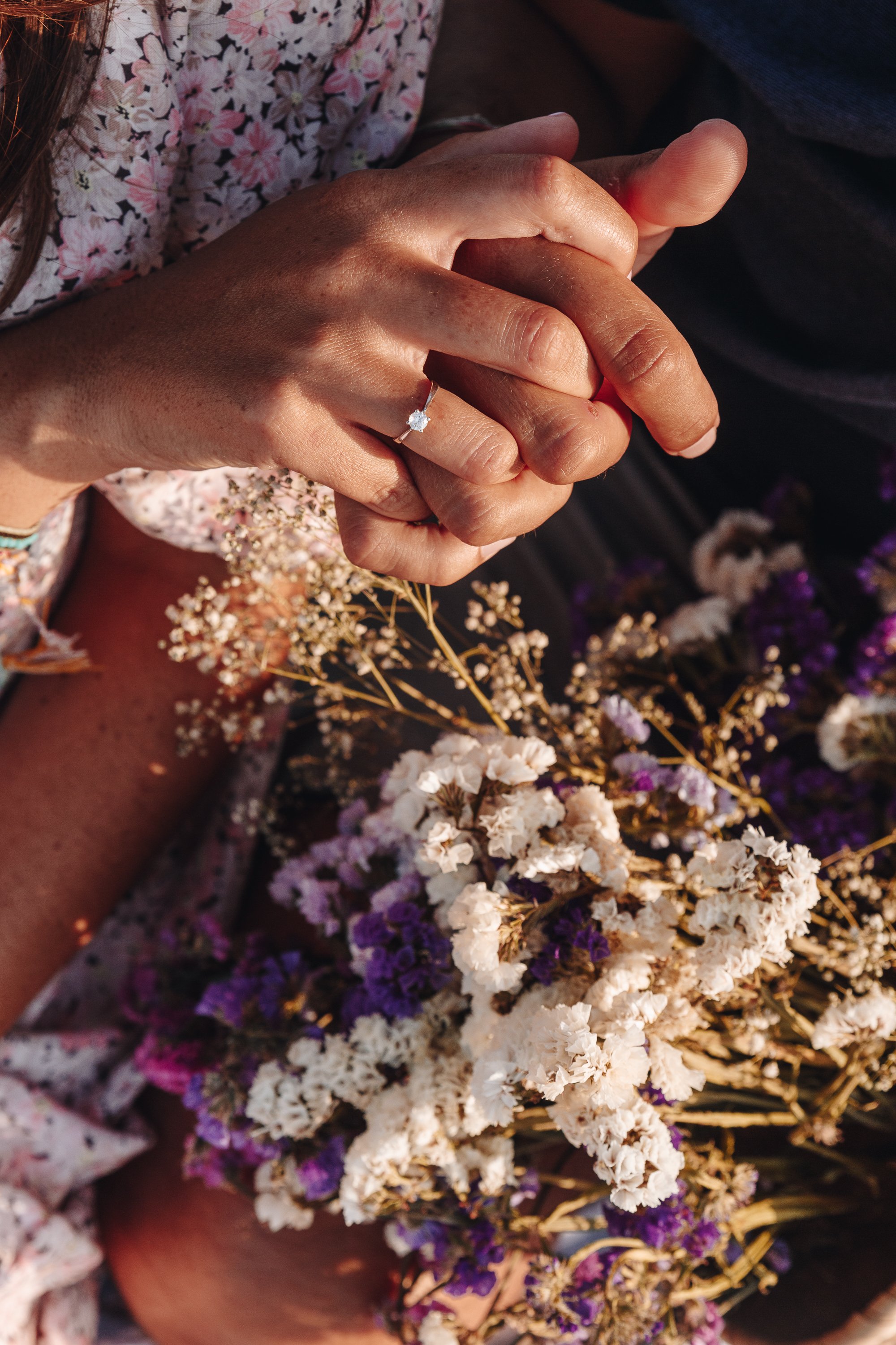 Close-up of the bride and groom’s hands with the engagement ring surrounded by flowers.