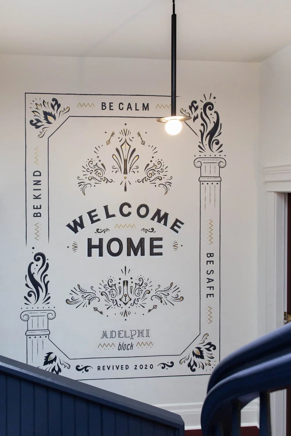 Decorative wall art with the phrases, "Be Calm," "Be Kind," "Be Safe," and "Revived 2020" surrounding a central message that reads, "Welcome Home." The design includes ornamental flourishes, floral motifs, and classical column capitals, with a single