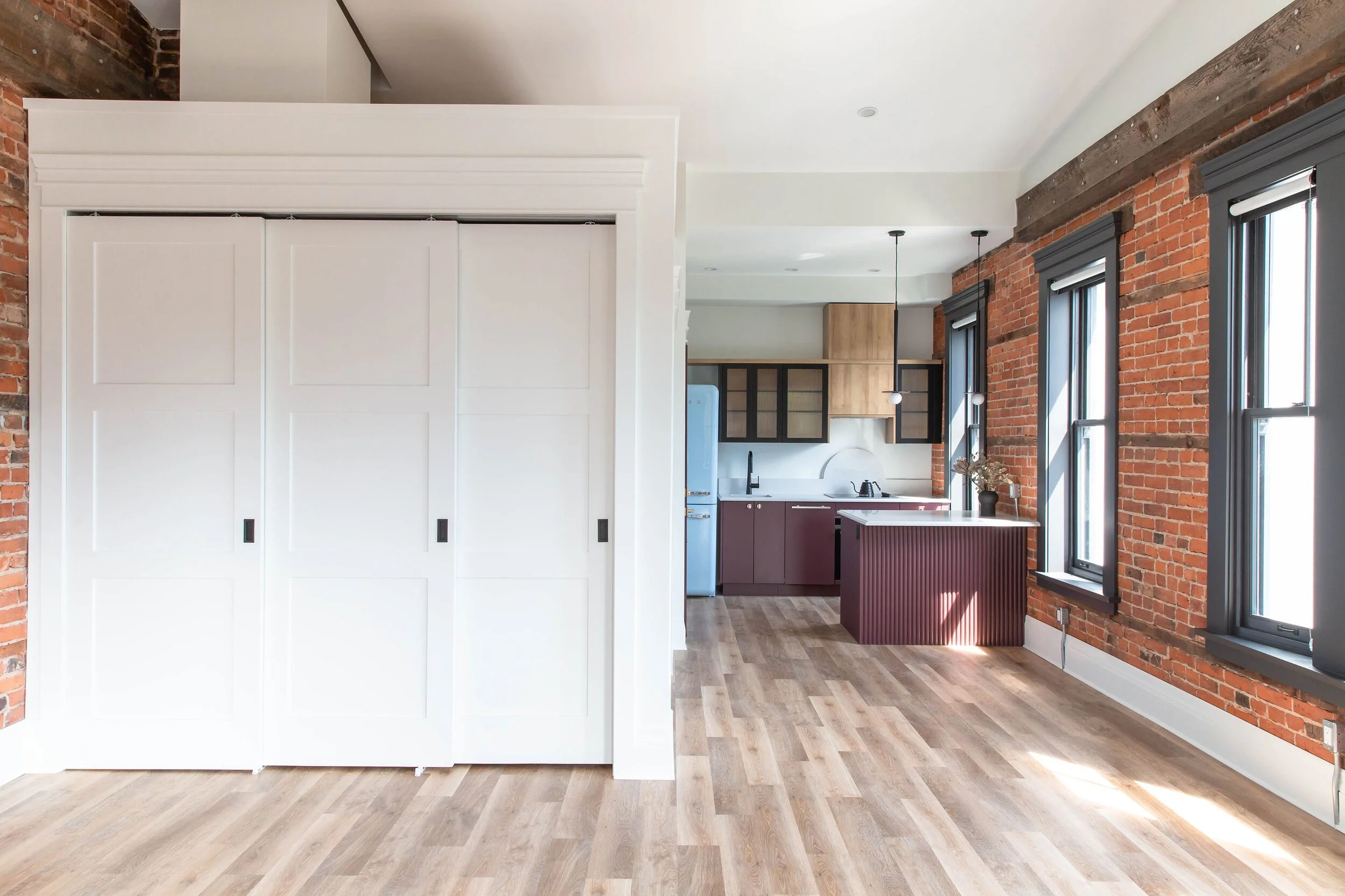 Open living space with exposed brick walls, large windows with black frames, wood flooring, and a view into a kitchen with purple lower cabinets, black upper cabinets, and light wood upper cabinets.