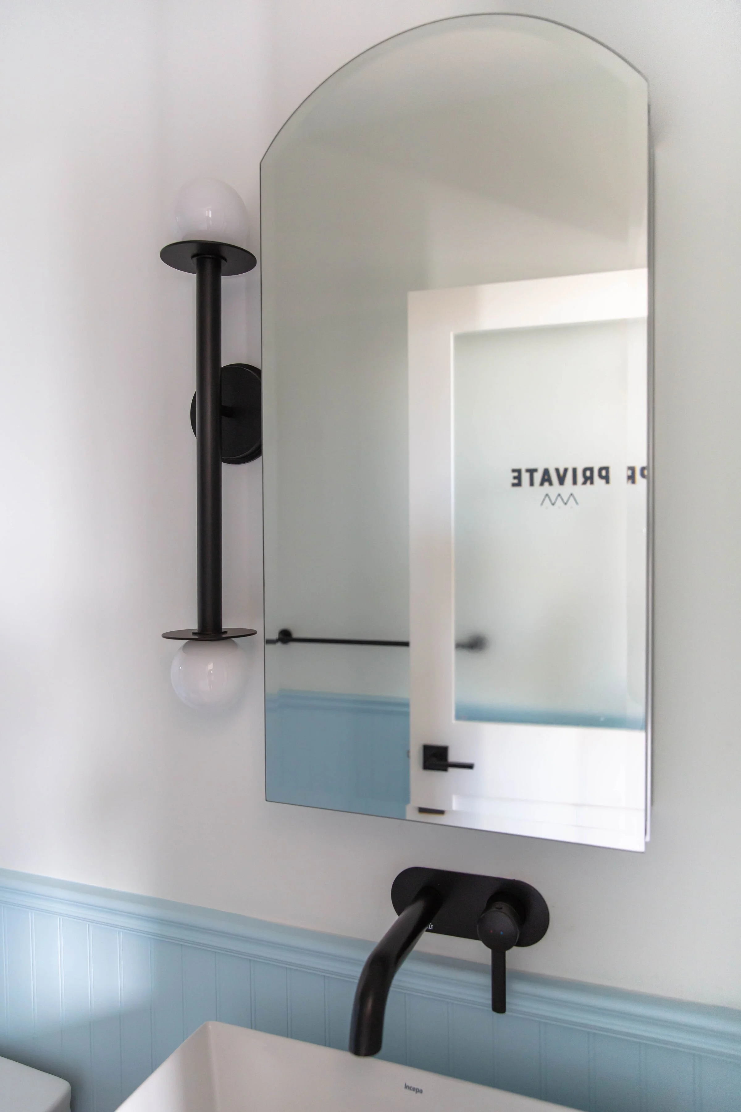 A bathroom mirror with a modern black wall mounted light fixture beside it, a white sink below, and a light-colored wall with light blue paneling.