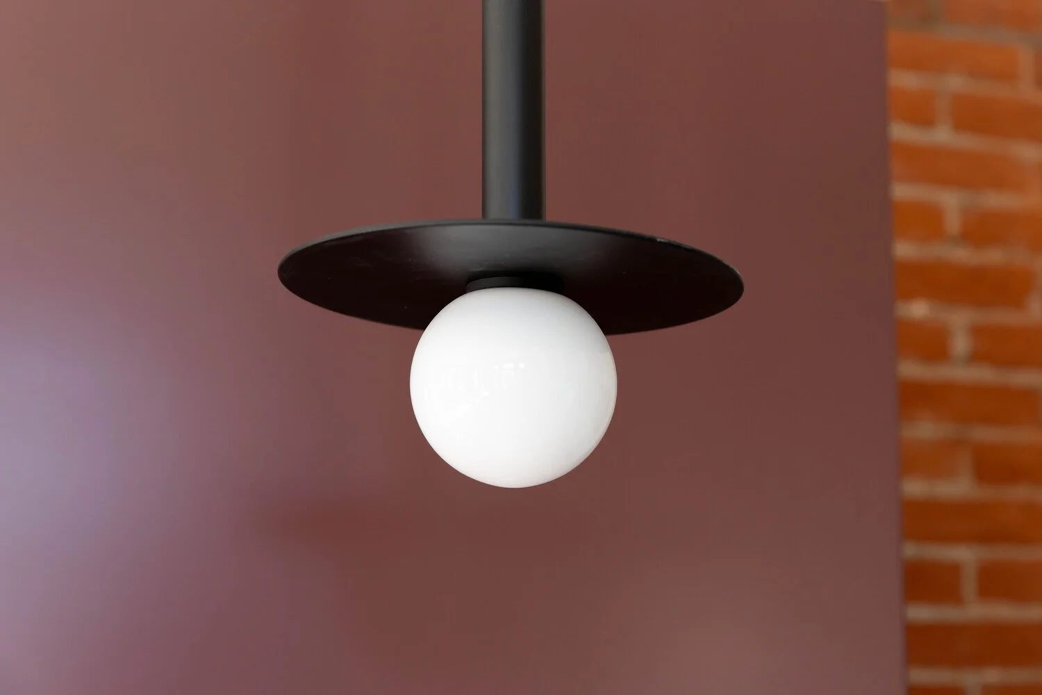 Close-up of a modern ceiling lamp with a black circular shade and a white spherical bulb against a pinkish wall and a brick wall in the background.
