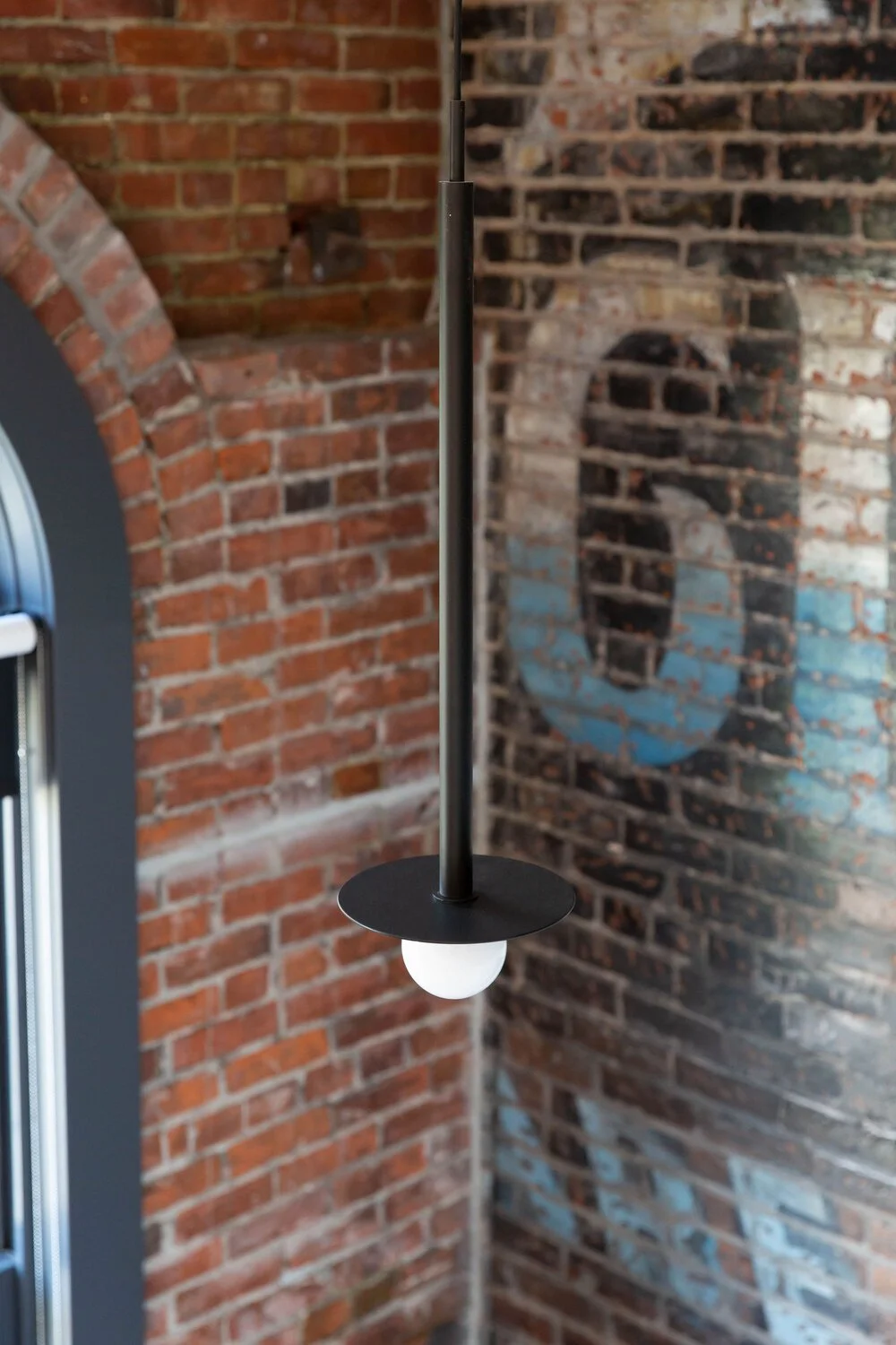 Wall-mounted lighting fixture with a white bulb and black circular shade mounted on a black pole on a brick wall background.