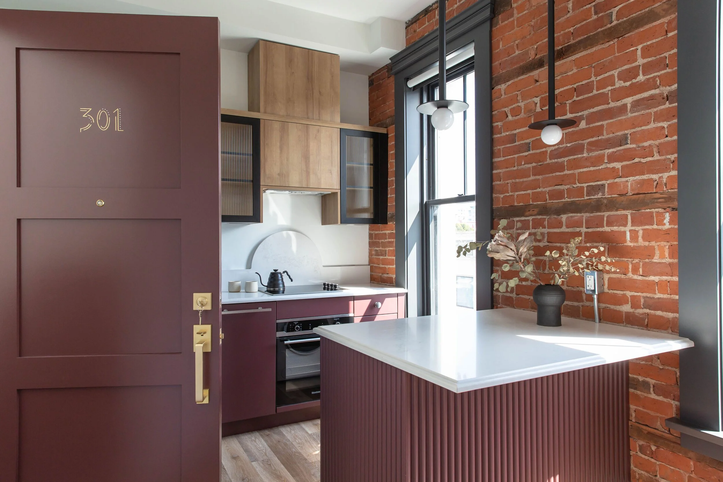 Interior of a modern kitchen with a brick wall, a purple door with room number 301, wooden and black cabinets, a white countertop, a black kettle, and a black vase with dried leaves near a window.