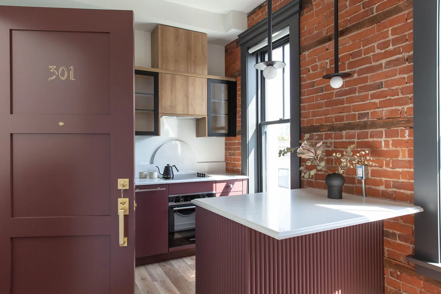Interior of a modern kitchen with exposed brick wall, wooden shelves, black and mauve cabinetry, a large window, and a white countertop with a black vase holding dried eucalyptus branches.