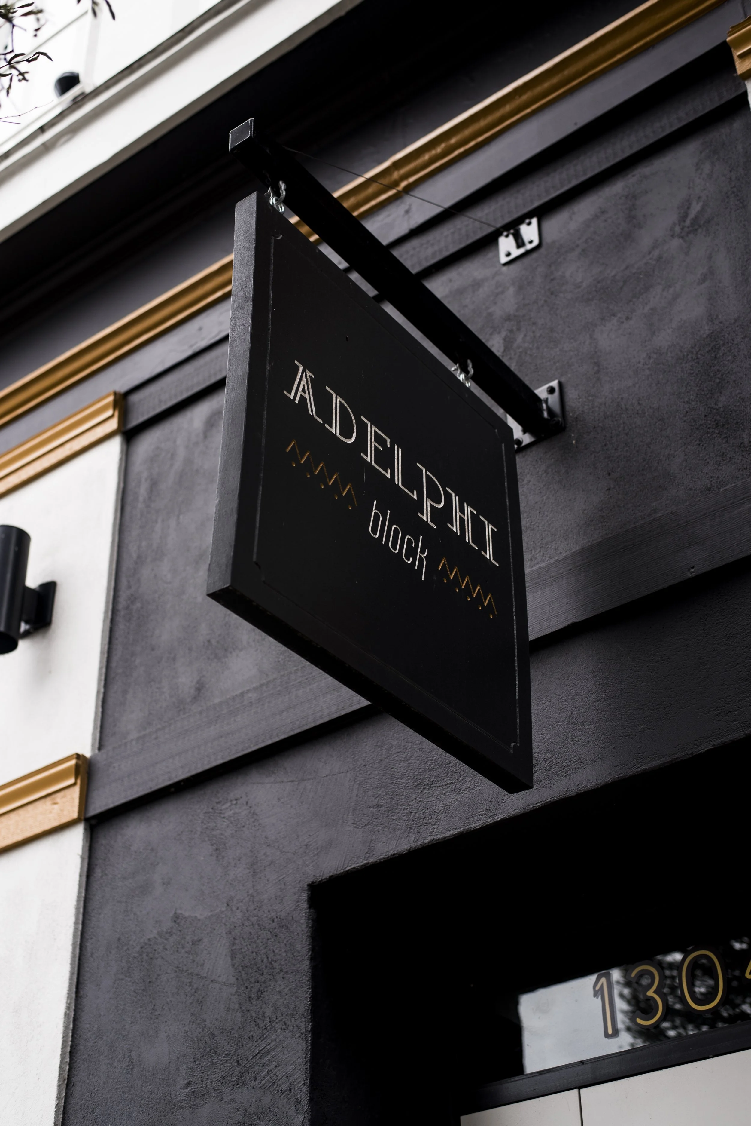 Black sign with gold and white text that reads 'JALDEELPHI block' hanging outside a building with dark gray and white walls and gold trim.