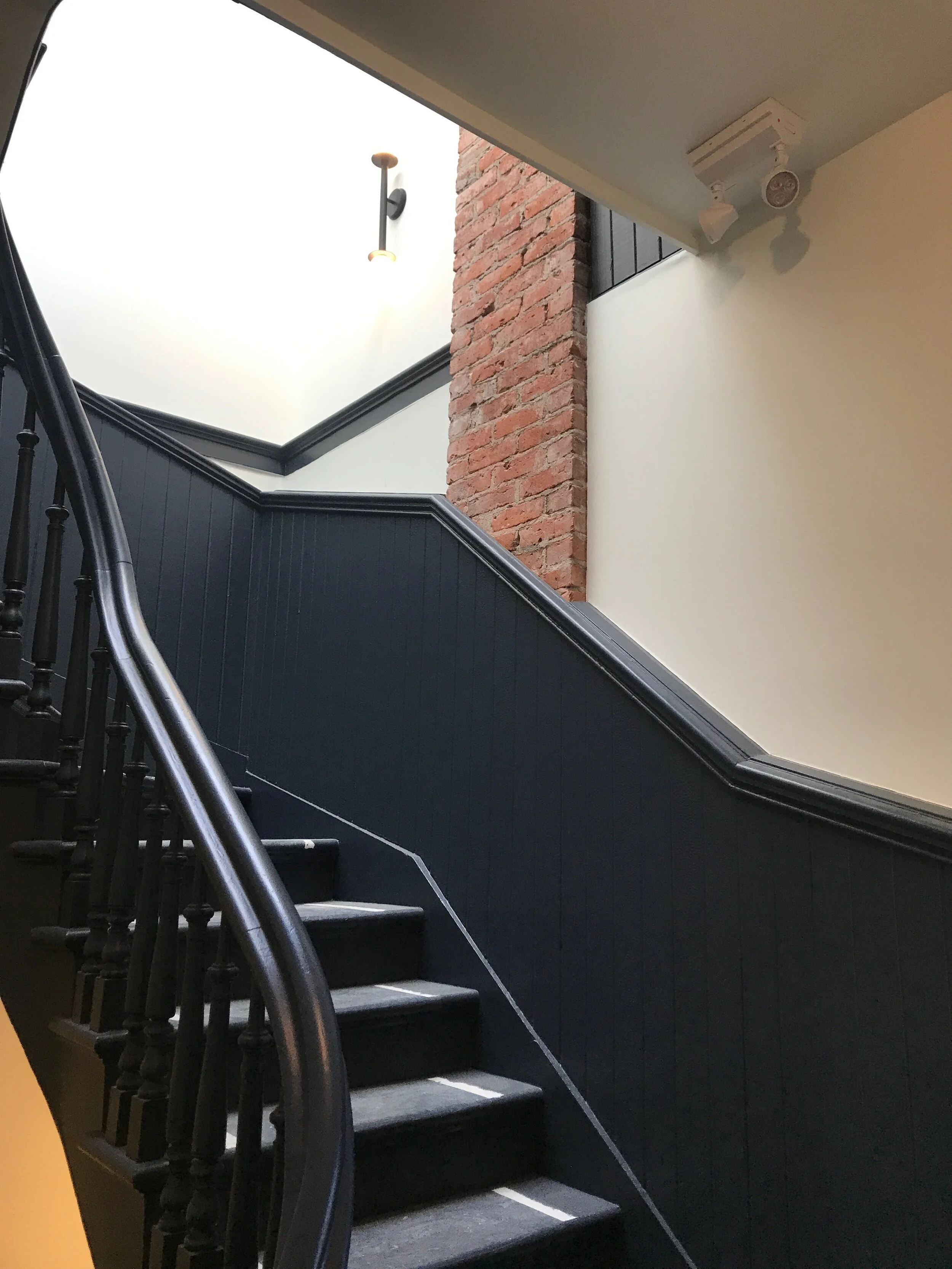 Interior staircase with black wooden handrails and steps, a dark wood-paneled wall, a red brick wall, ceiling light fixture, and a ceiling-mounted security camera.