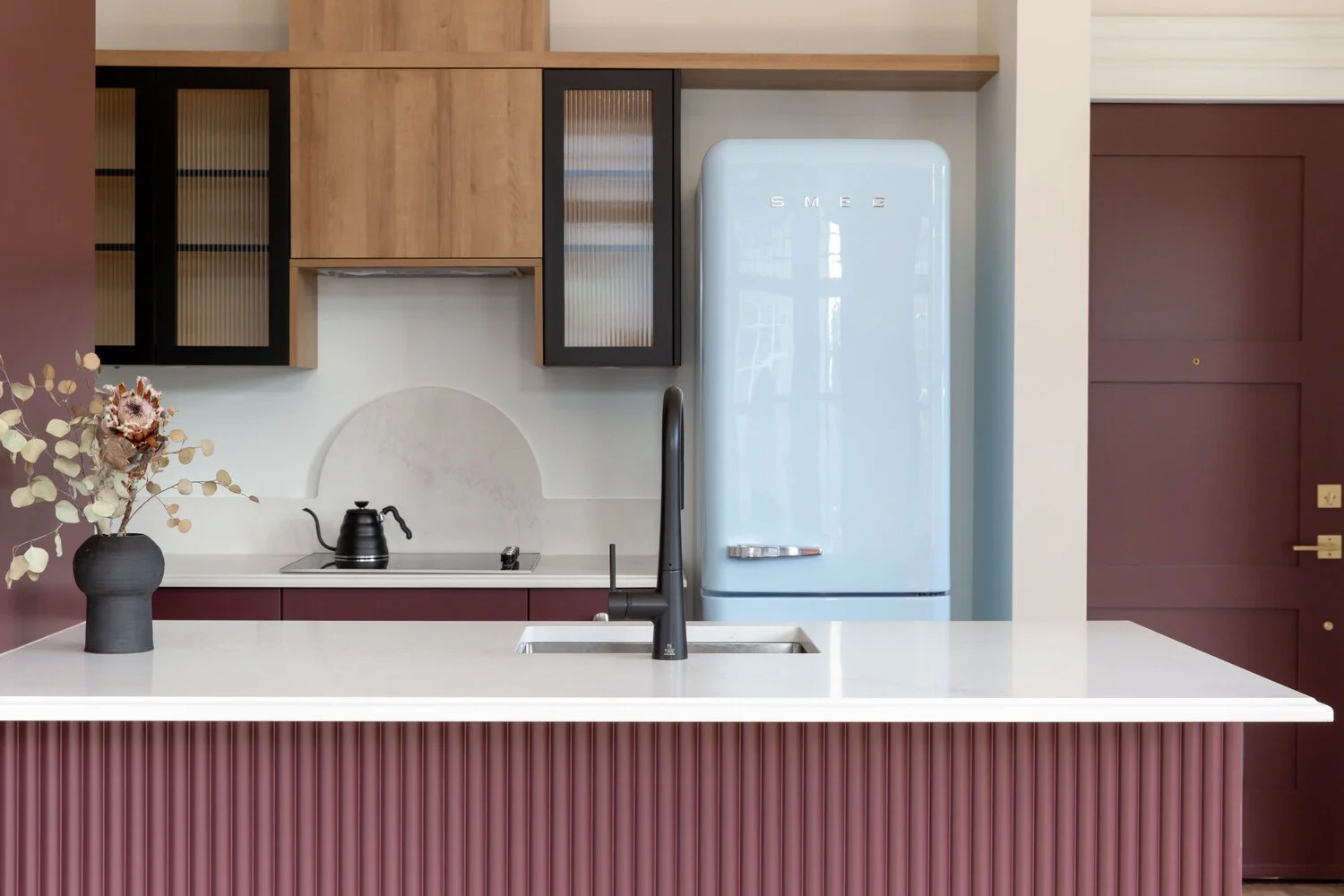 Modern kitchen with maroon island, black faucet, black kettle, light blue SMEG refrigerator, wooden and black cabinets, decorative vase with dried flowers, and purple door.