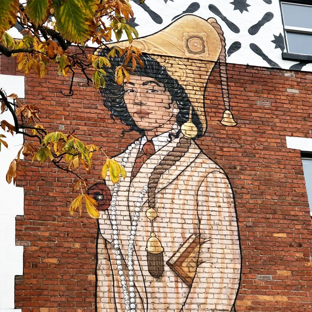 Mural of a woman with curly dark hair, wearing a wide-brimmed hat with bells, pearl necklace, and holding a small book, painted on a brick wall.
