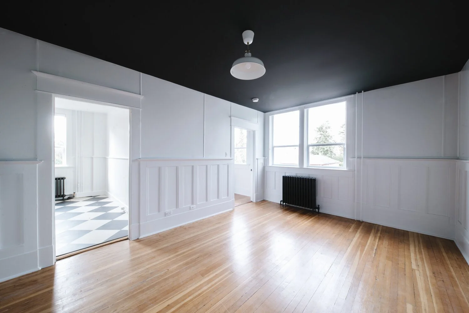 Empty living room with hardwood floors, white walls, large windows, and a black radiator, with an adjacent room visible through an open doorway.
