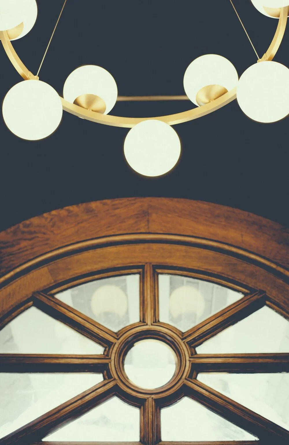 A modern chandelier with circular light bulbs hanging from a dark ceiling above a decorative wooden window with multiple panes.
