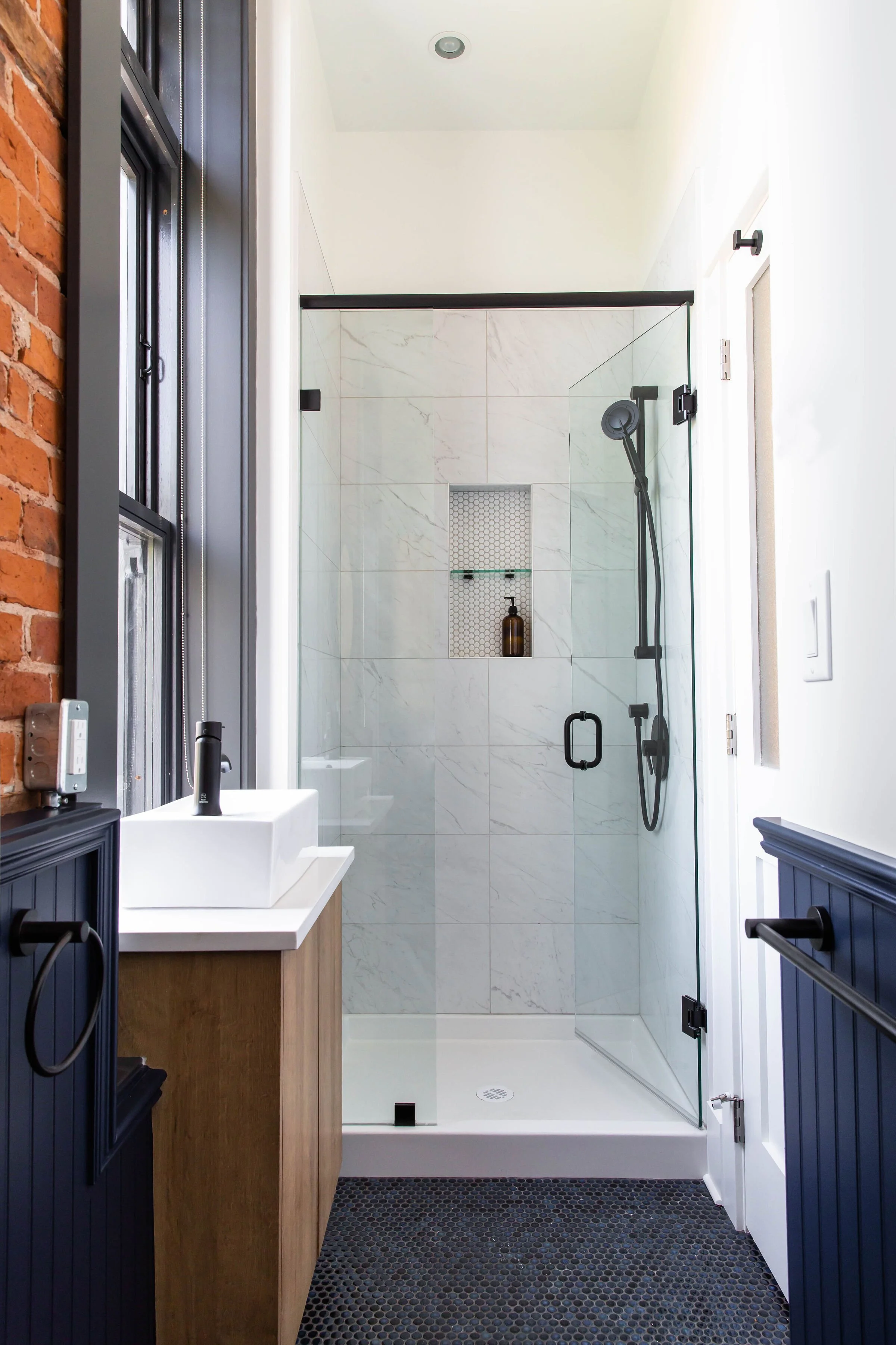 Modern bathroom with a glass-enclosed shower, white marble tiles, a built-in shower niche with a bottle, a small white sink on a wooden vanity, and a black textured floor mat.