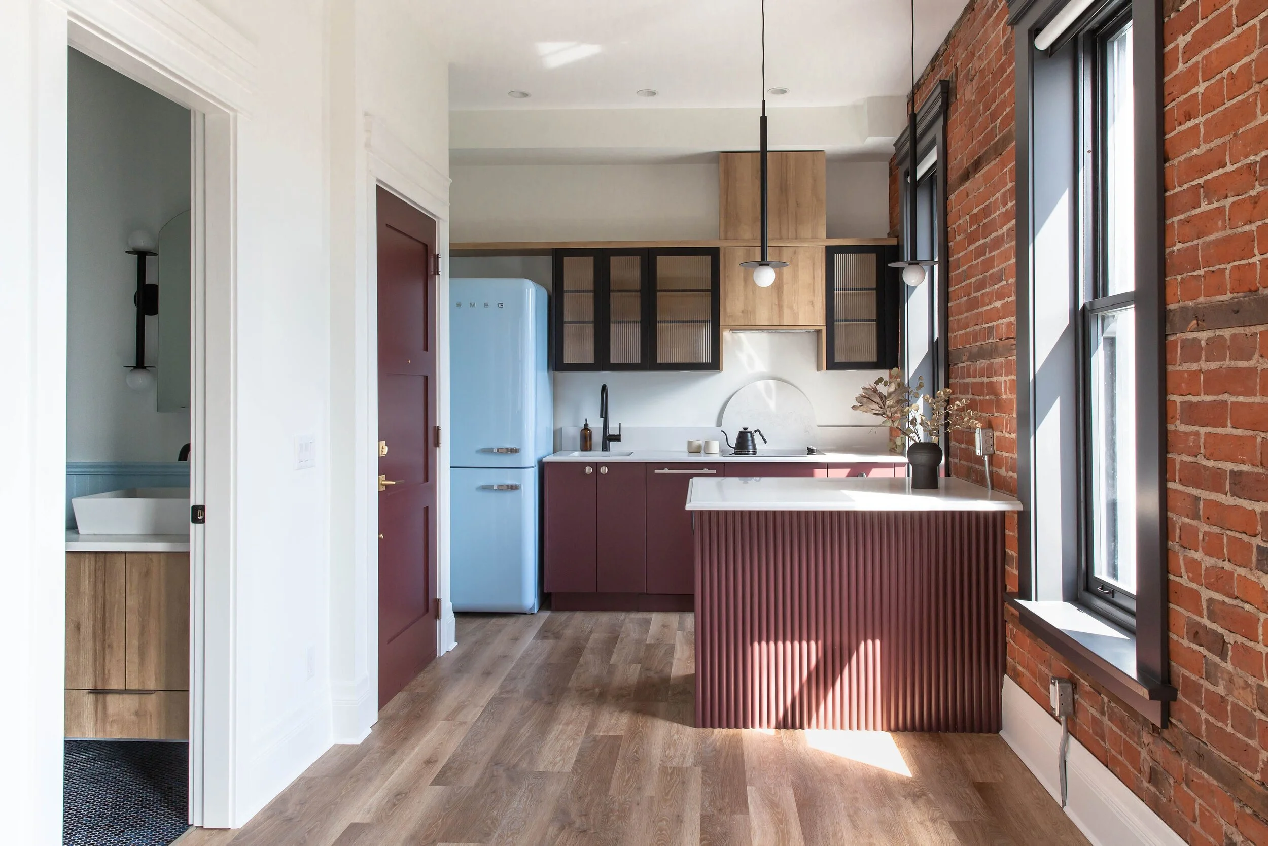 Modern kitchen with red brick accent wall, black and wood cabinetry, blue retro refrigerator, and pendant lighting over a white countertop island.