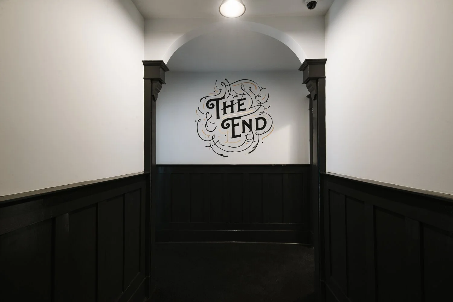 A hallway with white walls and black wainscoting, ending in an arch with a sign that reads 'The End' surrounded by decorative swirls.