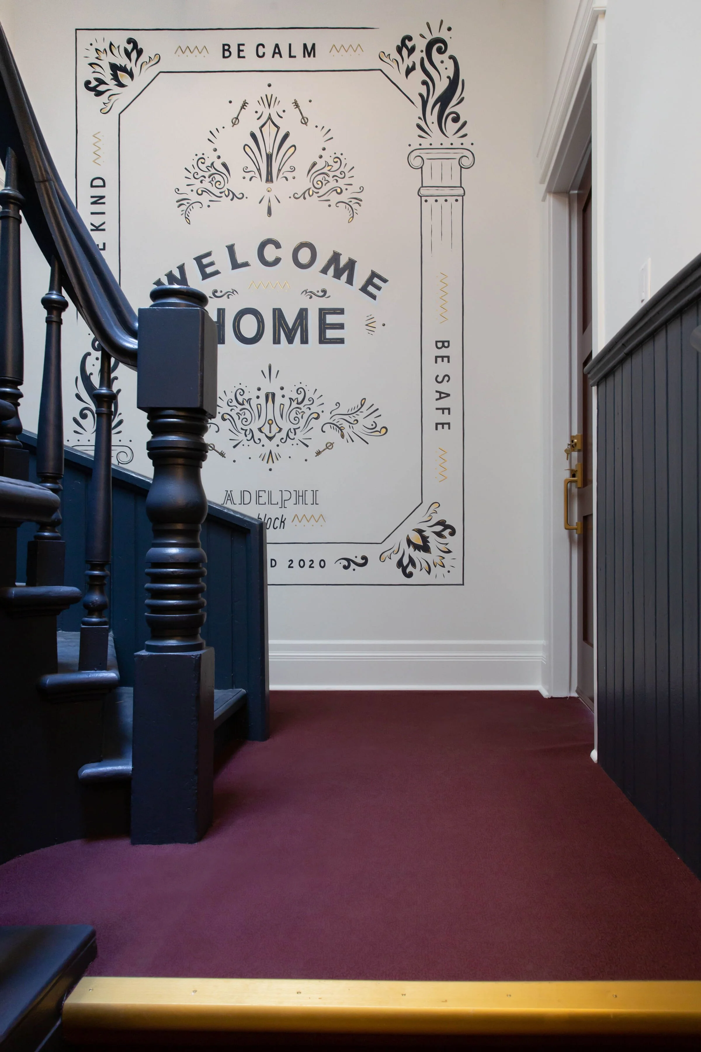 Interior of a home with a dark wood staircase on the left and dark wood paneling on the right. A large decorative wall sign saying "Welcome Home" is at the back, with black and gold accents and phrases like "Be Calm," "Be Safe," and "Be Kind" around