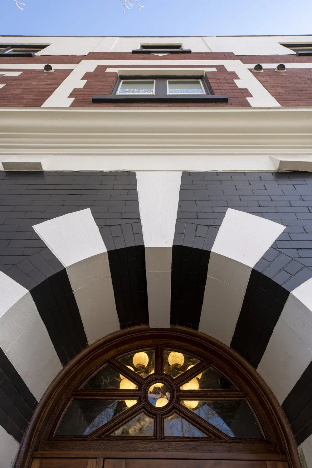 Close-up of an ornate arched doorway with black and white striped pattern, hanging lights visible through a circular window, part of a historic brick building.