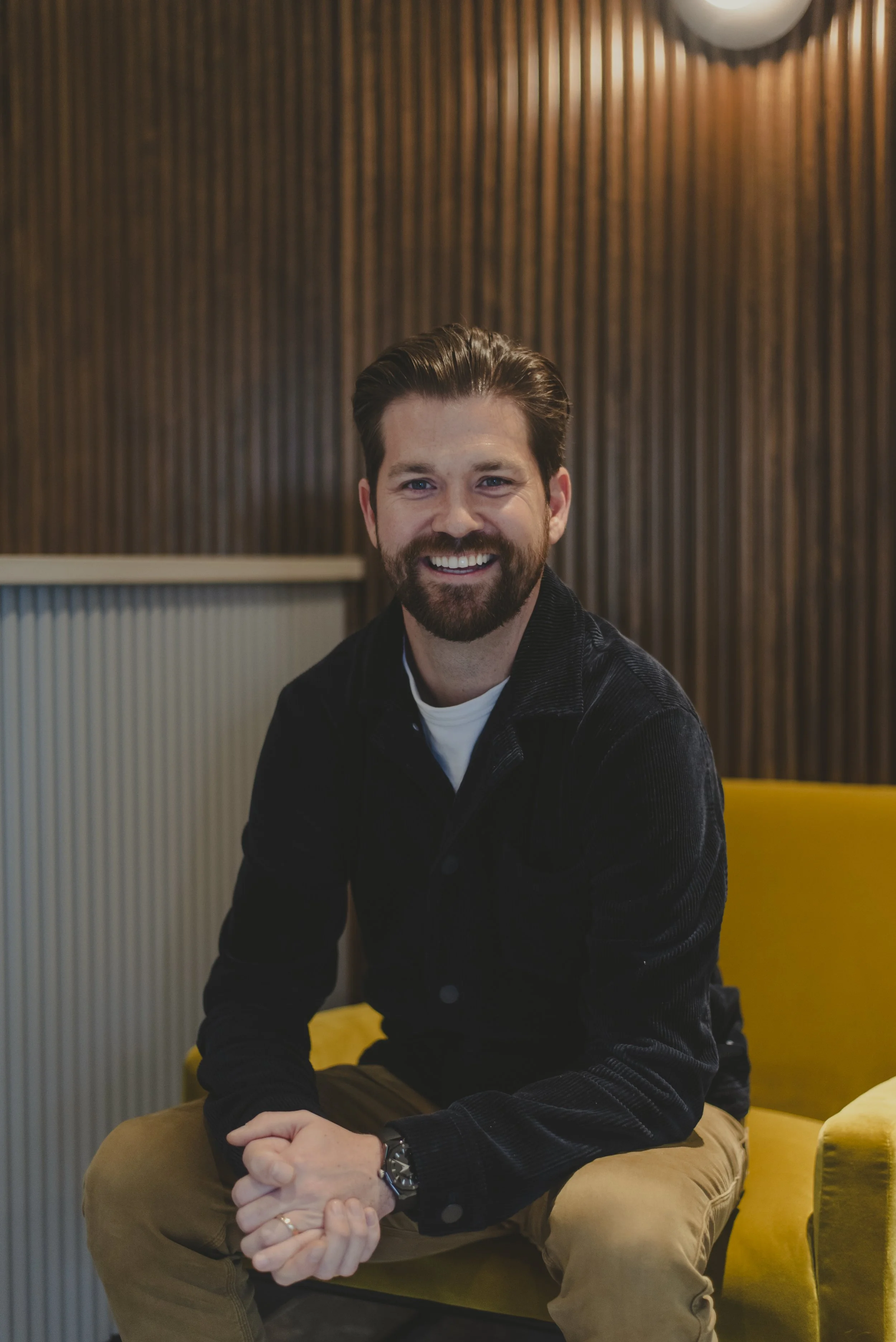 A man with a beard and mustache smiling, standing with arms crossed in an office or commercial space, with a dark wood wall behind him featuring a backlit sign that reads '9TC'.