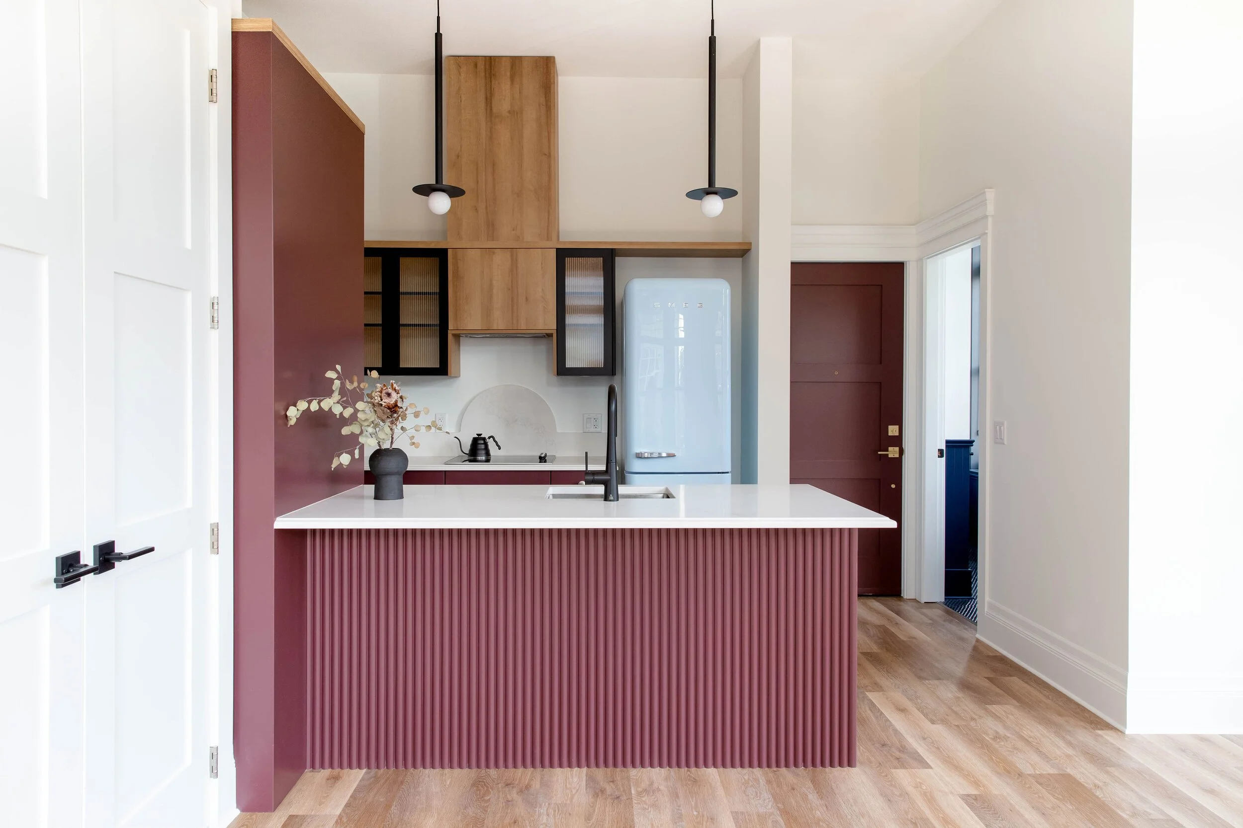 Modern kitchen with maroon island and wooden cabinets, white countertops, black faucet, black pendant lights, vintage-style refrigerator, and a vase with dried flowers.