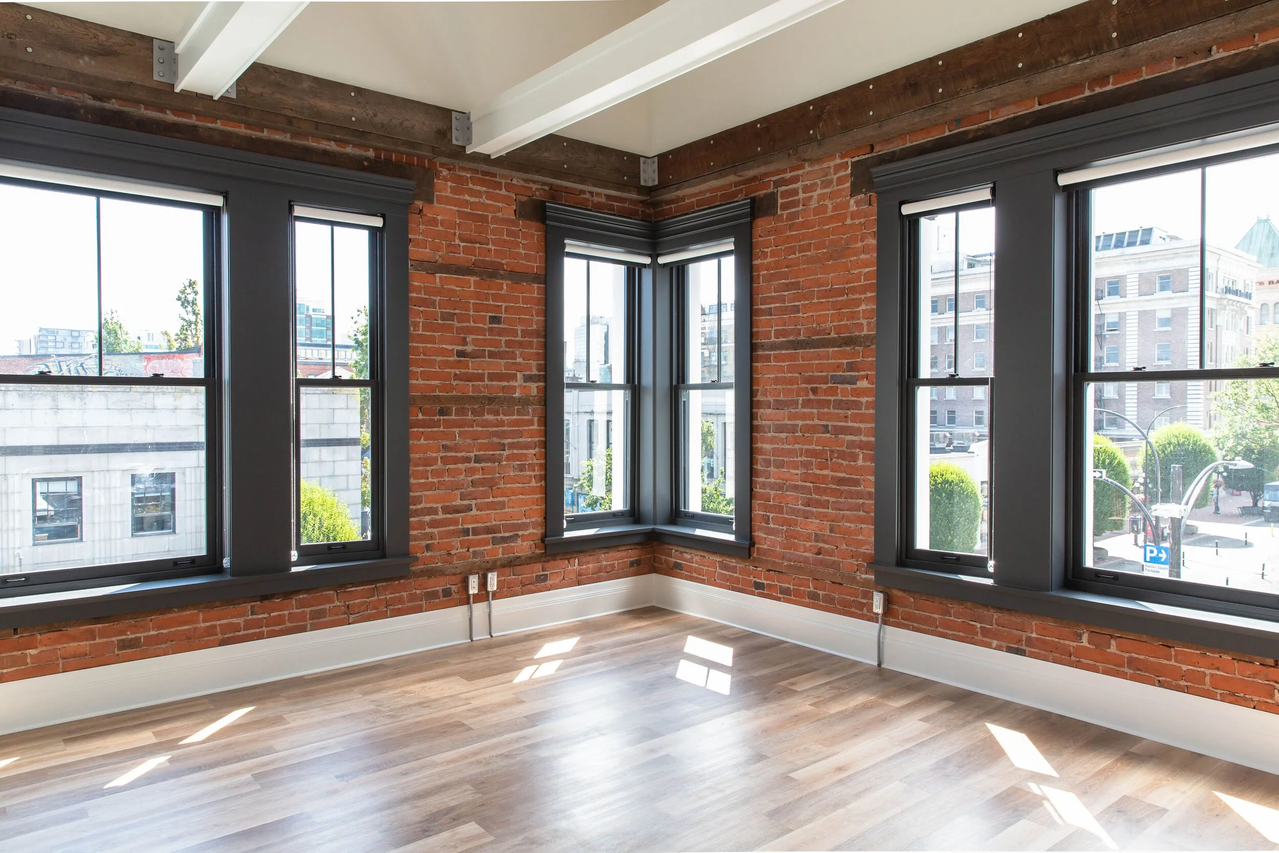 Empty room with large windows, exposed brick walls, light-colored wood flooring, and white ceiling beams.