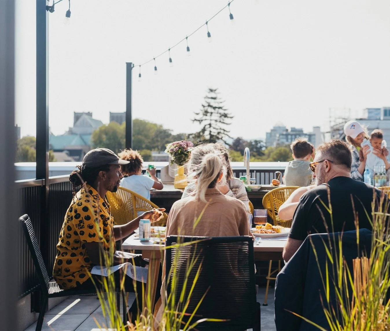 People dining outdoors on a rooftop terrace during daytime, with string lights hanging overhead, city skyline in the background, and a variety of food and drinks on the tables.
