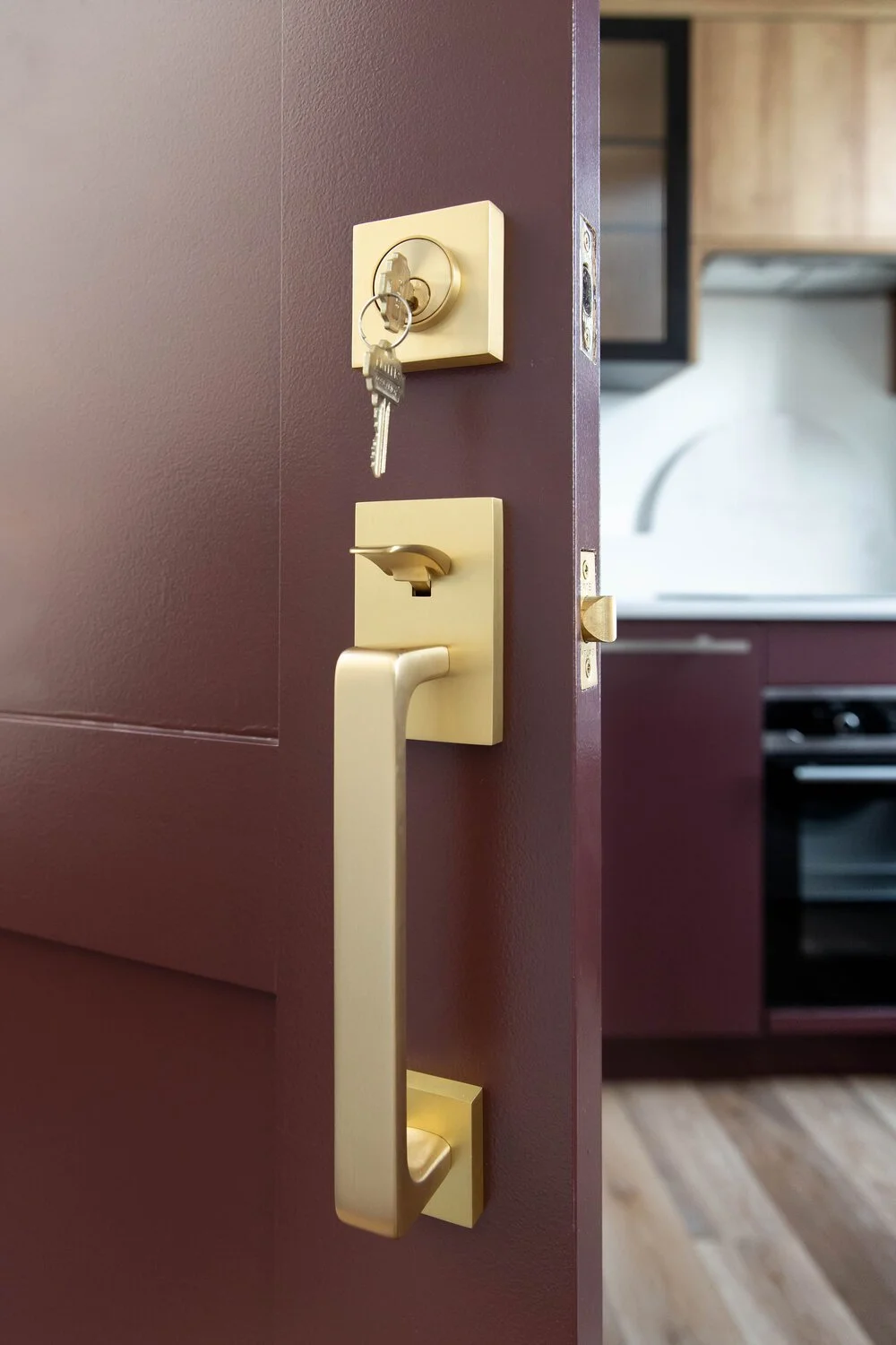 Close-up of a lock with keys hanging on a door with a brass handle, leading into a kitchen area.