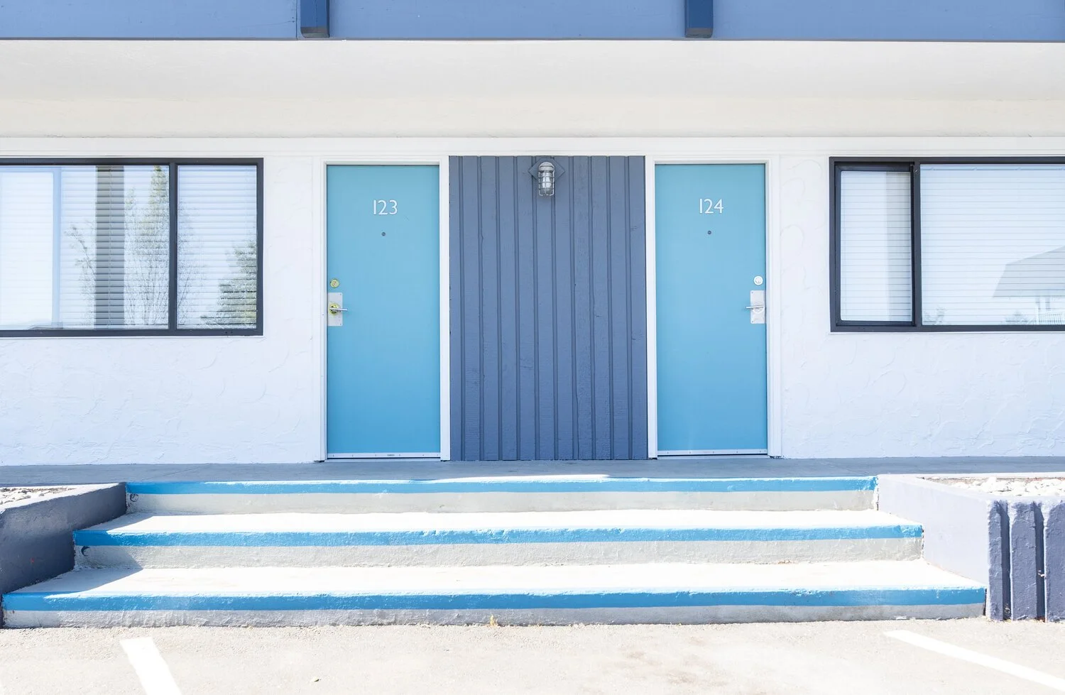 Exterior view of a motel or apartment building with two blue doors numbered 123 and 124, black-framed windows with blinds, a concrete staircase, and a blue wall above.
