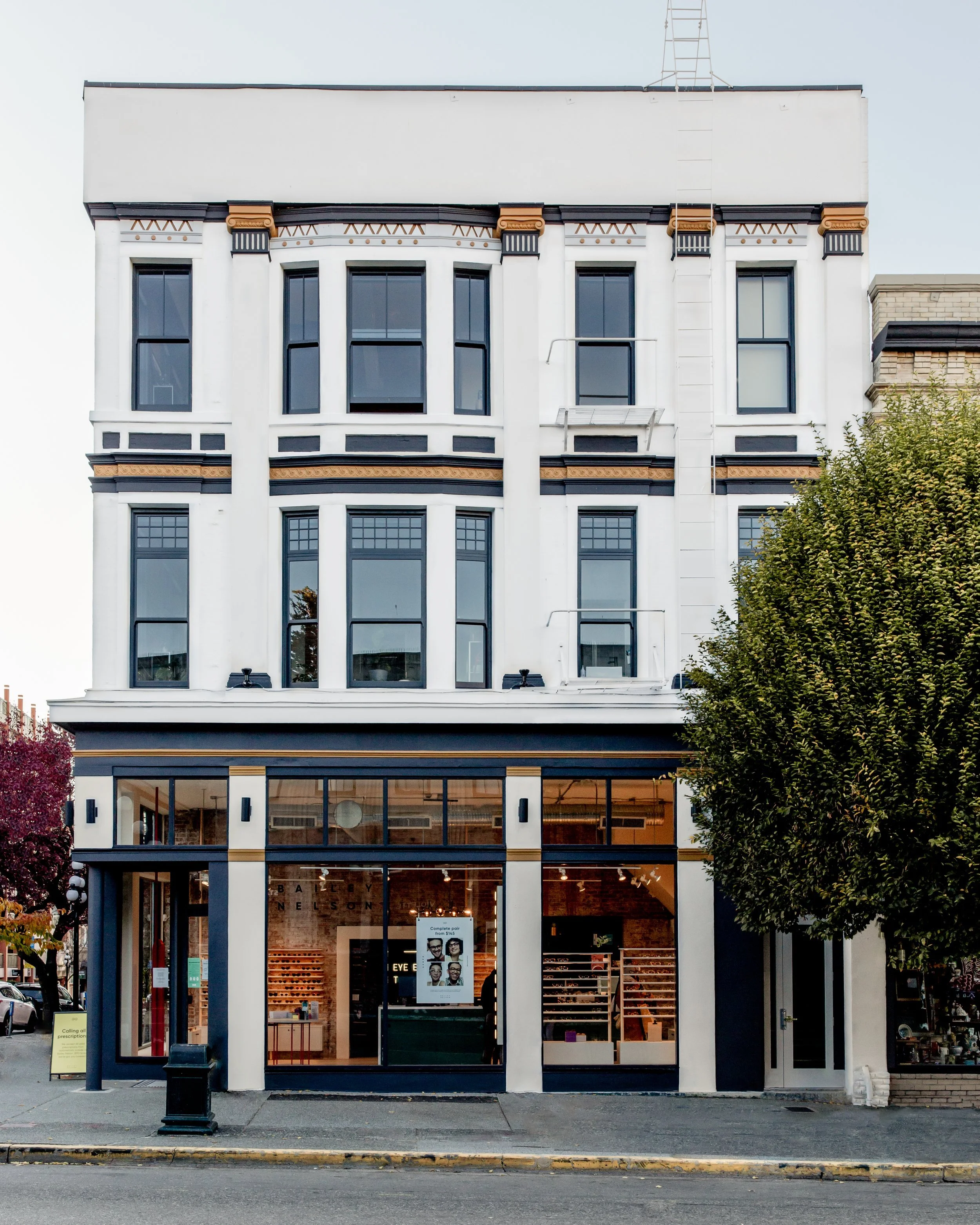 Front view of a multi-story white building with large ground floor windows, displaying a shop interior, and upper floors with black framed windows and decorative trim, with trees on both sides and a ladder on the roof.