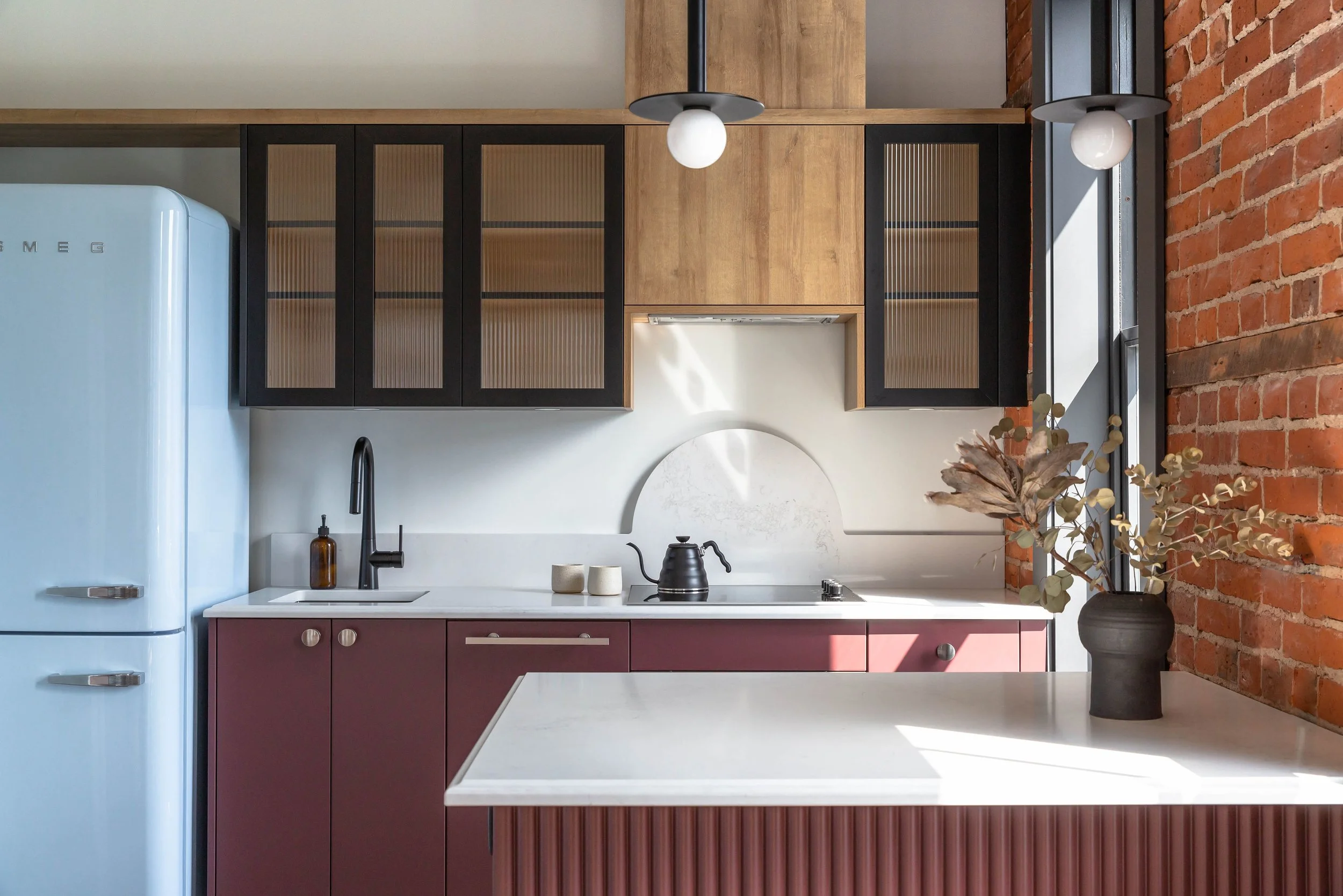 Modern kitchen with white marble countertop, burgundy lower cabinets, black upper cabinets with glass doors, exposed brick wall, black kettle, black faucet, and black vase with dried flowers.