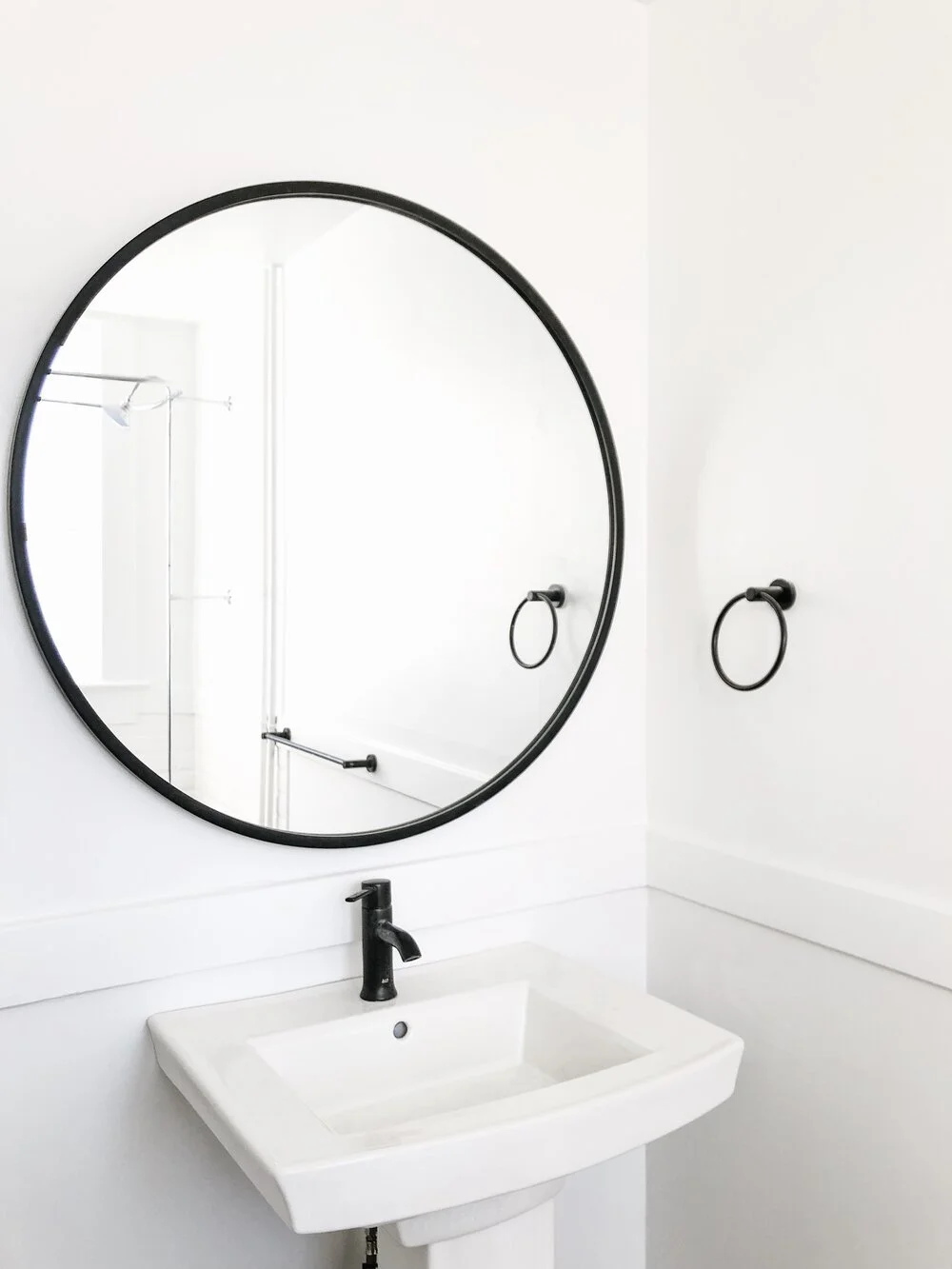 Bathroom with a white sink, black faucet, large round mirror, and black towel rings.