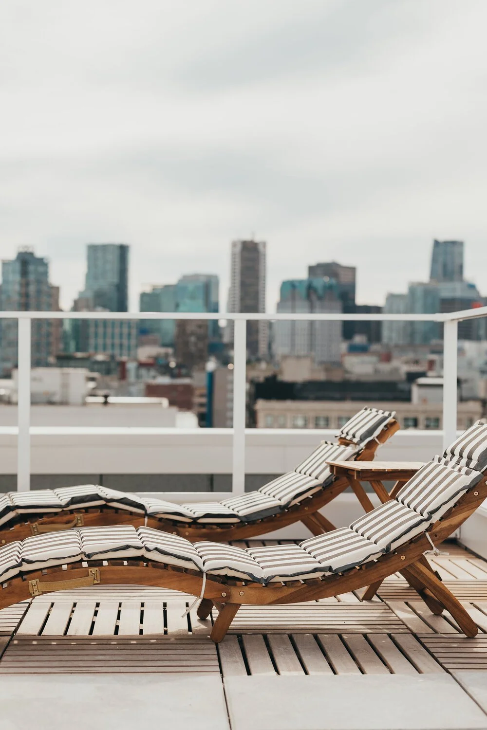 Two wooden lounge chairs with striped cushions on a rooftop terrace overlooking a city skyline with tall buildings under an overcast sky.