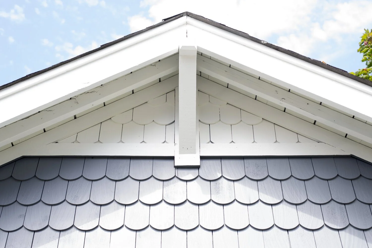 Close-up of a house's gabled roof showing white and gray decorative shingles and trim against a blue sky with clouds.