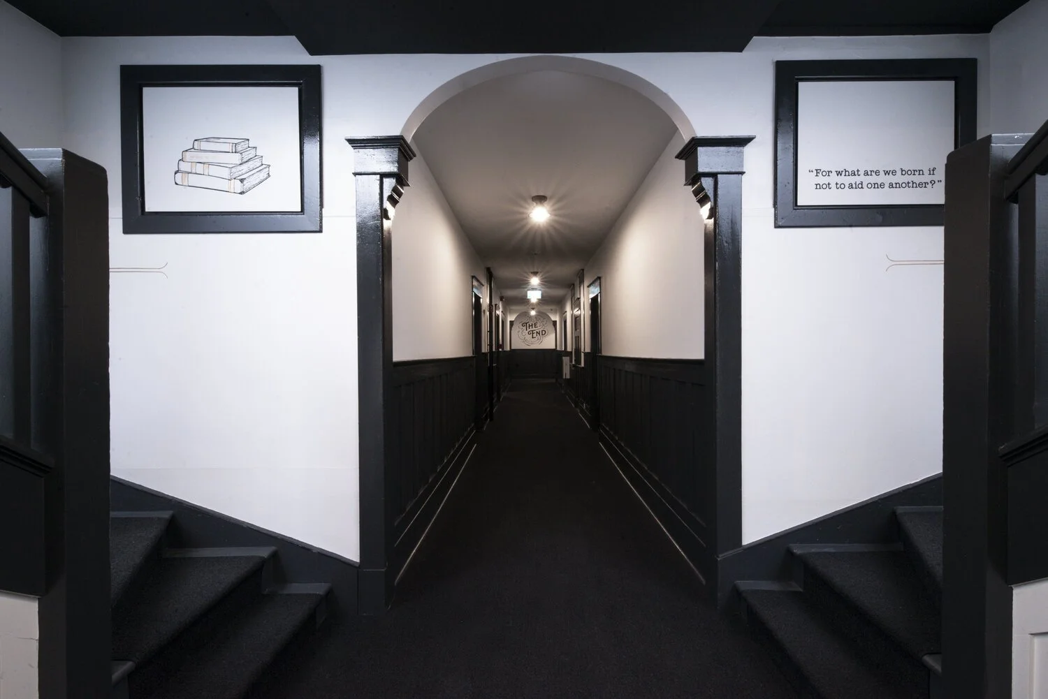 Long hallway with black wainscoting and white walls, framed artwork, and quote. The end of the hallway has a sign reading "The End."